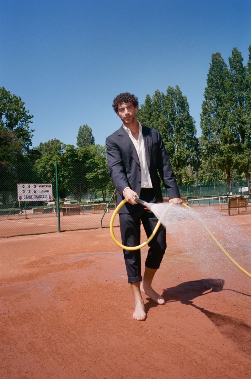 Man in suit with bare feet holds a yellow hose spraying water on a red clay tennis court with a blue sky and green trees in the background.