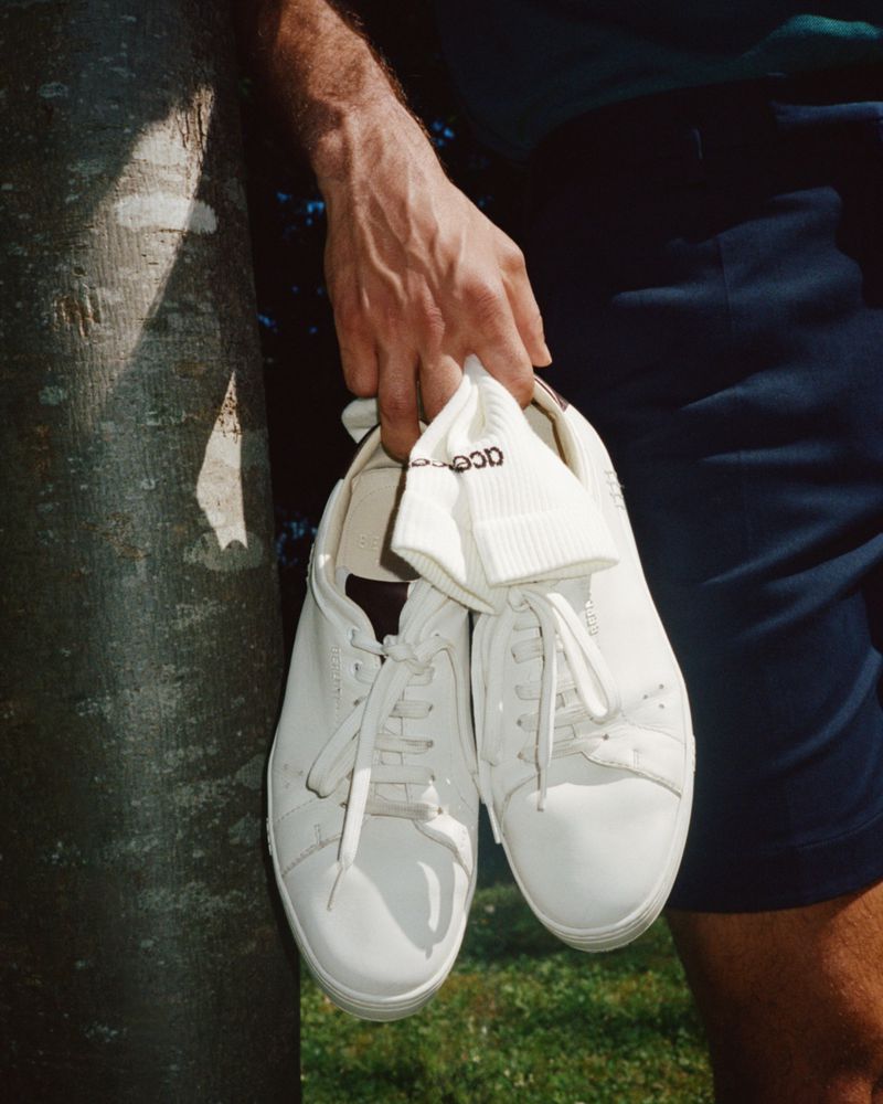 White leather sneakers are held in an outdoor setting next to a tree trunk with a glimpse of grass and a person's leg.