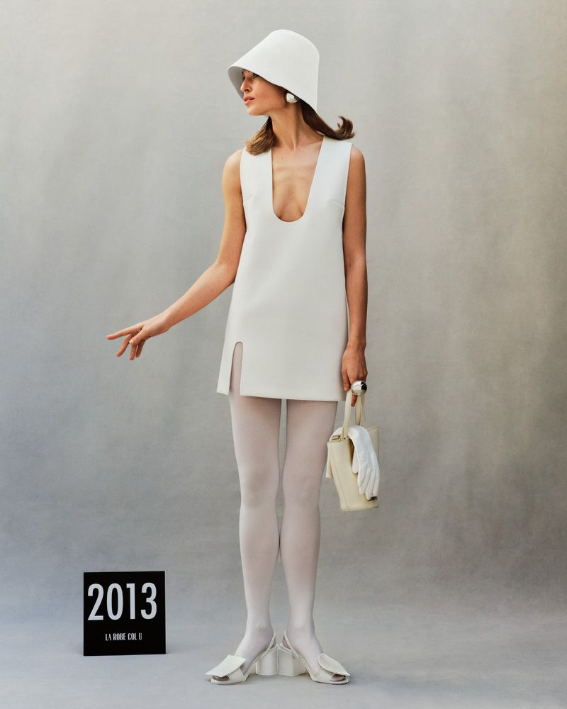 A woman in a white mini-dress and hat poses in front of a gray wall, holding a small white bag, showcasing 1960s-inspired fashion.