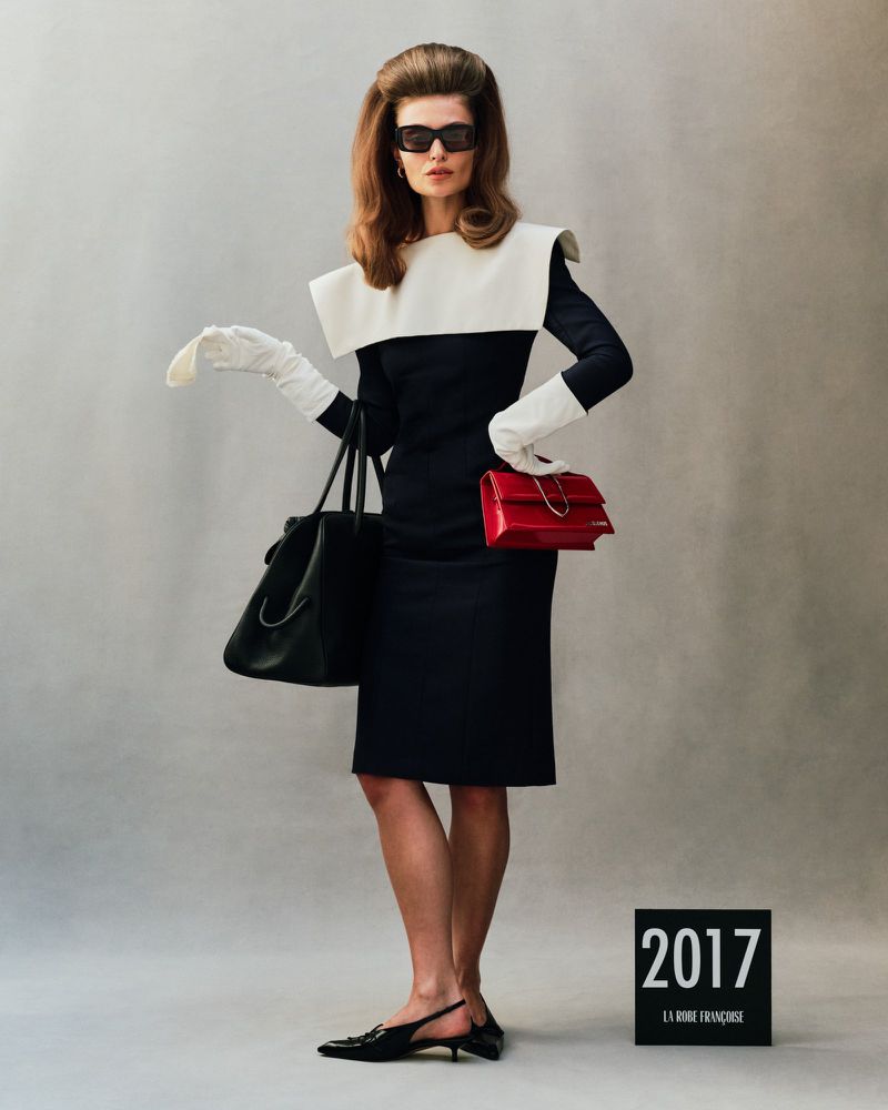 Elegant adult woman in a black and white dress, gloves, sunglasses, and accessories, posing in a studio with a gray backdrop.