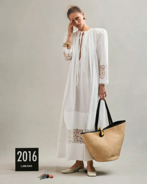 Fashion shot of a female model wearing a long white dress, holding a straw bag in a studio with keys and signage props.