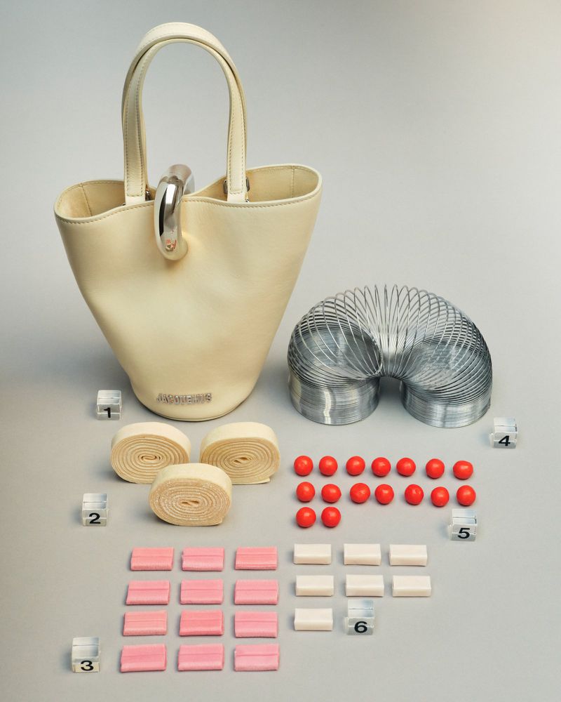 Ivory-colored purse with silver hardware is displayed next to a metal spring toy, candy and geometric cubes against a grey backdrop.