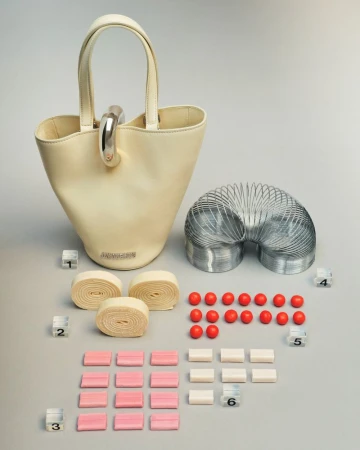 Ivory-colored purse with silver hardware is displayed next to a metal spring toy, candy and geometric cubes against a grey backdrop.
