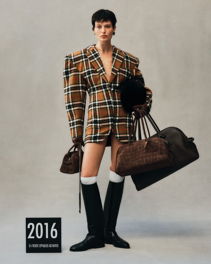 Fashion editorial shot of a woman in a plaid blazer dress, knee-high boots, and carrying handbags, set against a neutral backdrop.