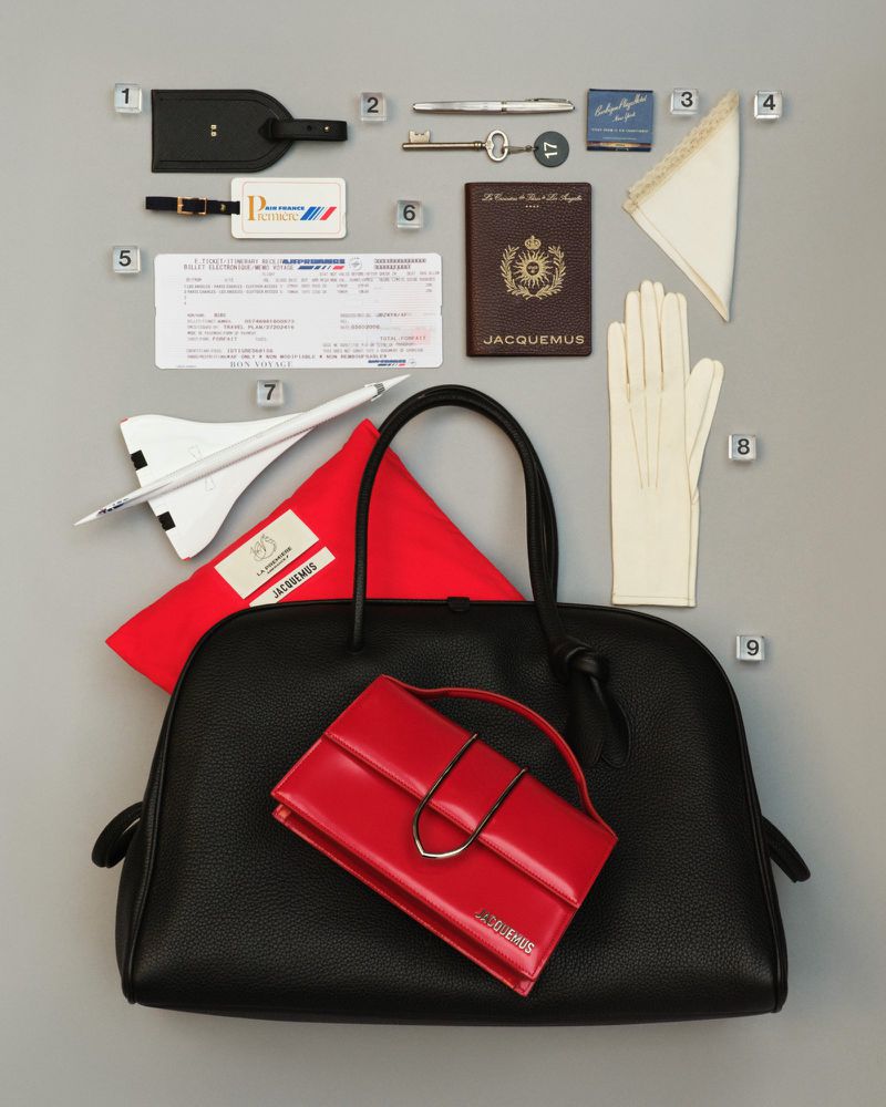 Overhead studio shot of black and red designer handbags, airplane model, and travel accessories, arranged on grey backdrop.