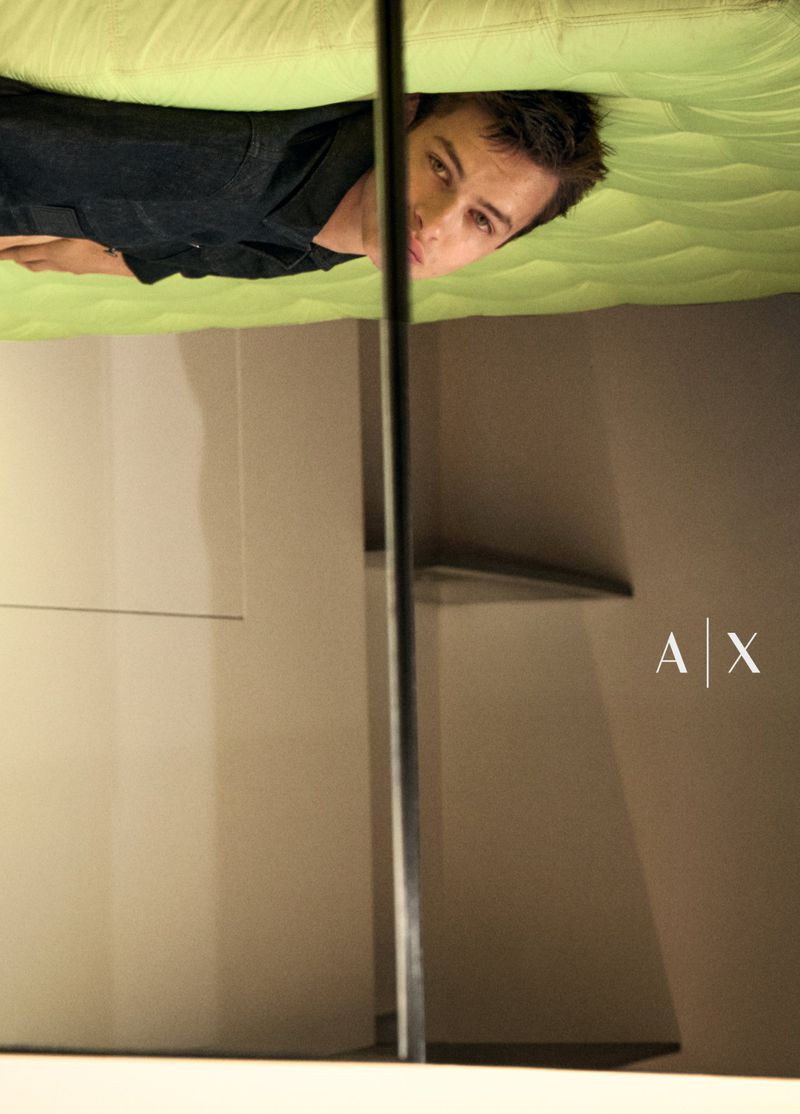 Portrait of a brunette man lying on a green bed, captured in an indoor setting with muted lighting and visible logo.