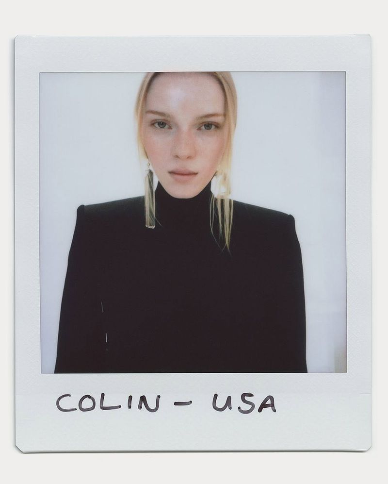 Portrait of a blonde woman with light skin wearing a black turtleneck sweater and long, clear earrings against a plain background.
