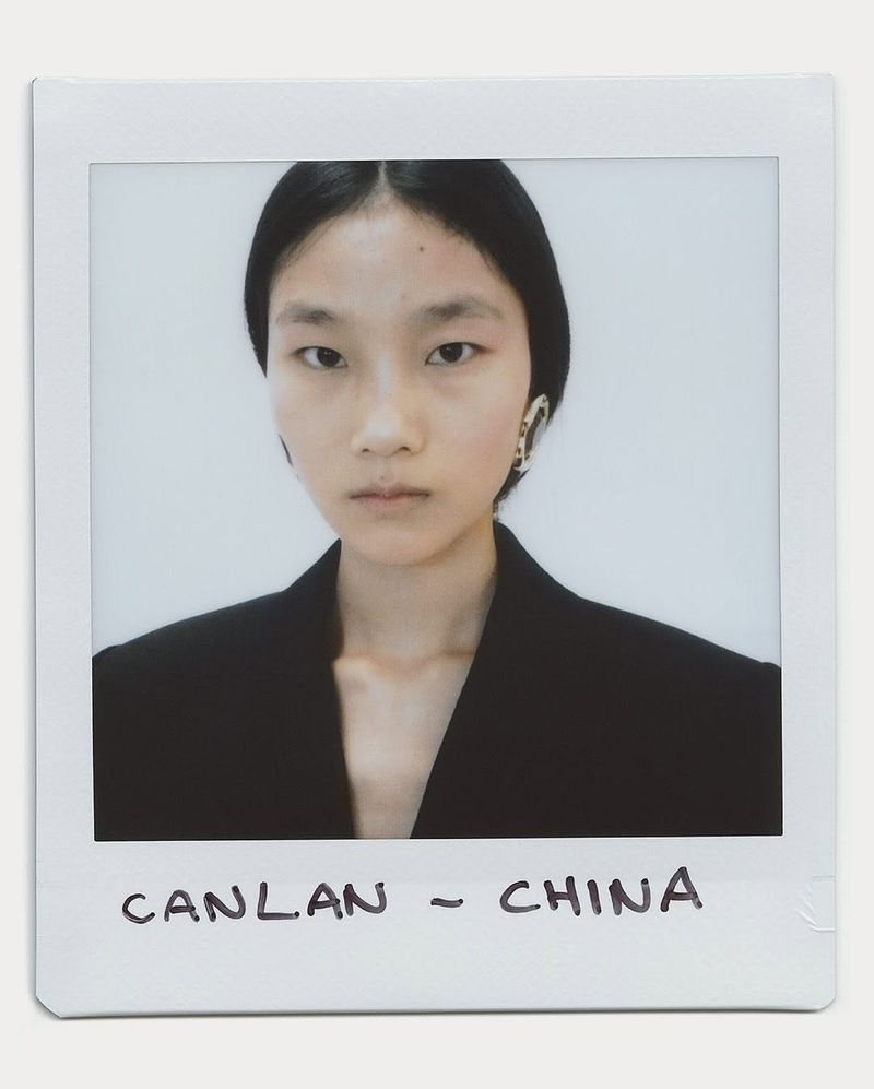 Close-up portrait of a young Asian woman with dark hair wearing a black jacket and earrings against a white background in a polaroid frame.