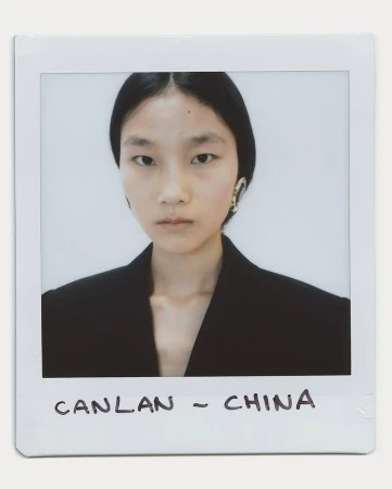 Close-up portrait of a young Asian woman with dark hair wearing a black jacket and earrings against a white background in a polaroid frame.