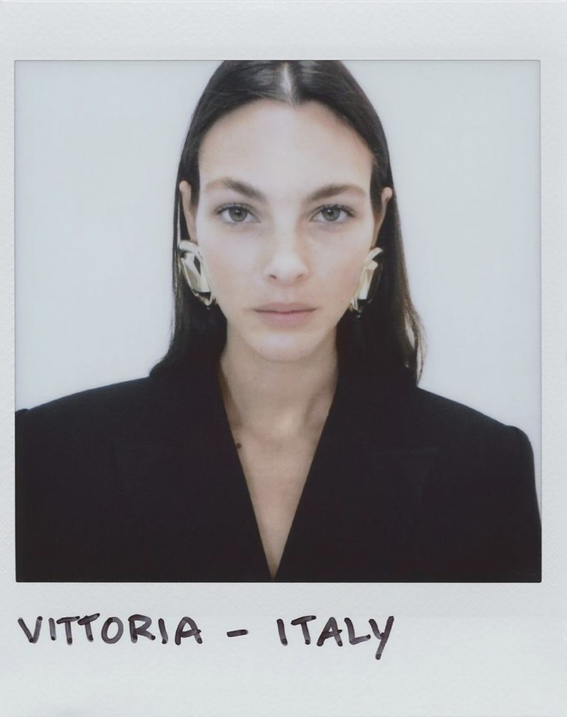 Close-up studio portrait of a brunette woman wearing large metal earrings and a black blazer against a white backdrop in a polaroid frame.