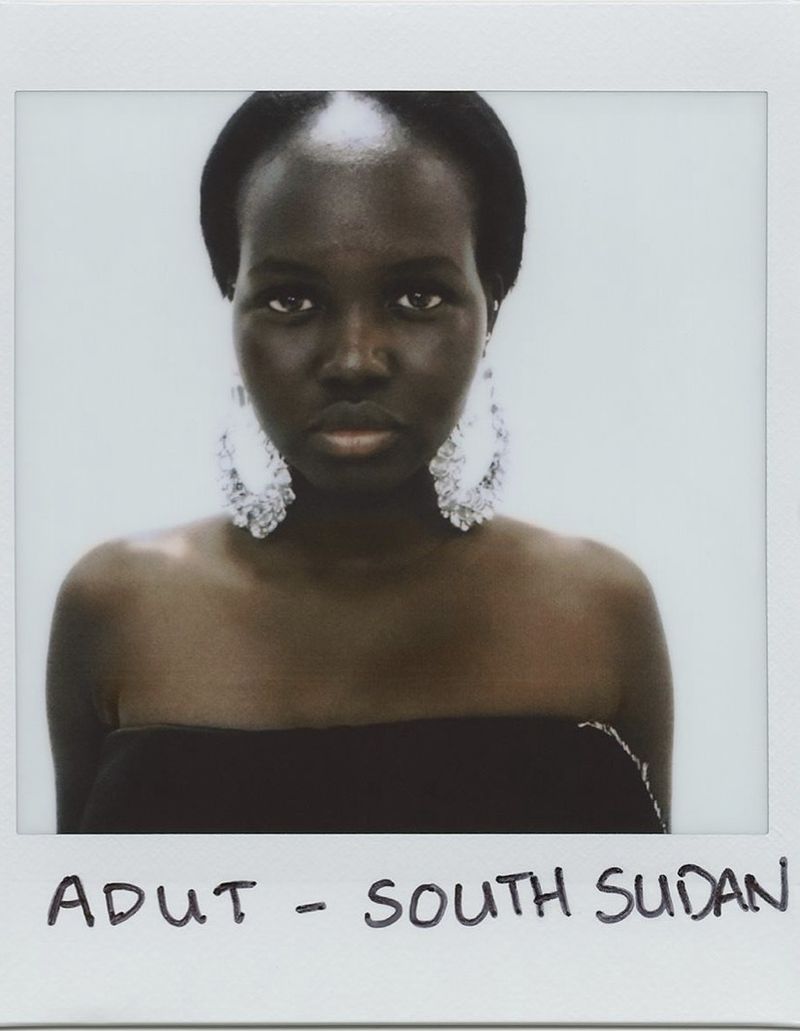 Close-up portrait of a Black woman with large, ornate earrings against a white background, framed within a white Polaroid border.