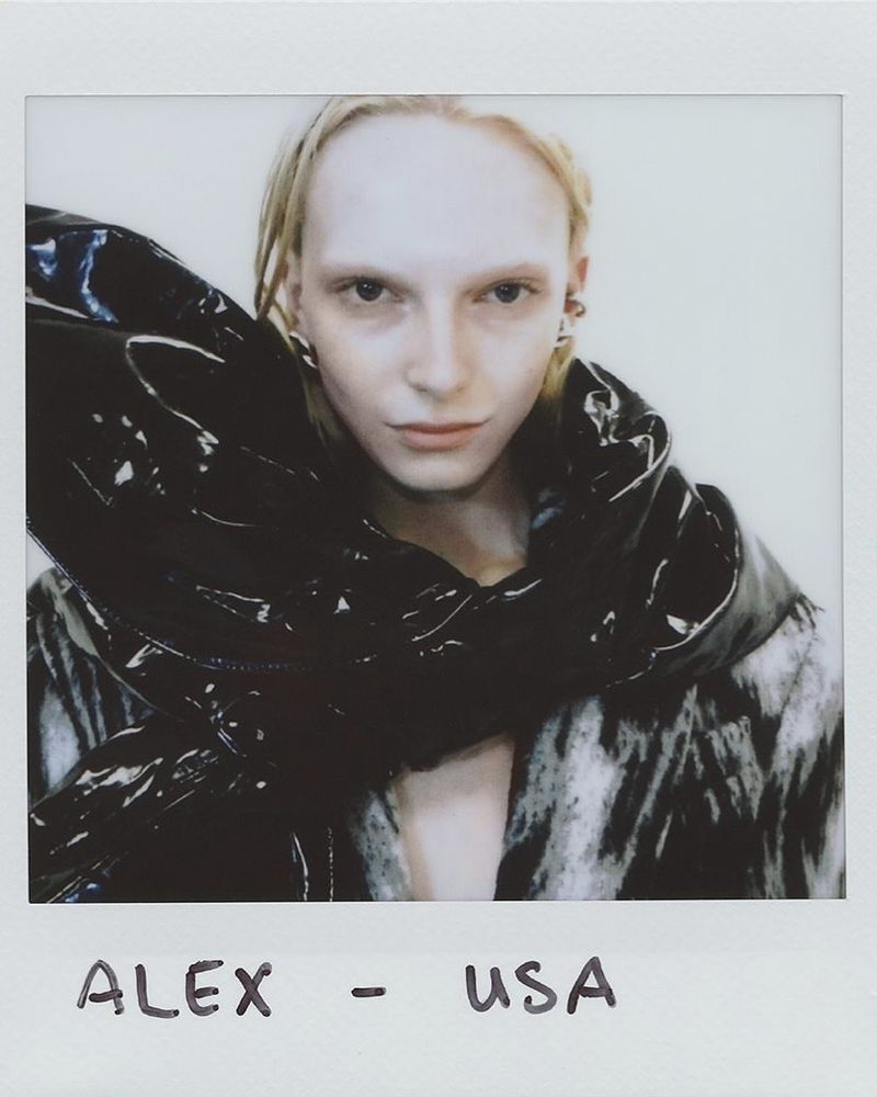 Close-up portrait of an adult woman with pale skin, blonde hair, and a unique black patent leather garment in a studio environment.
