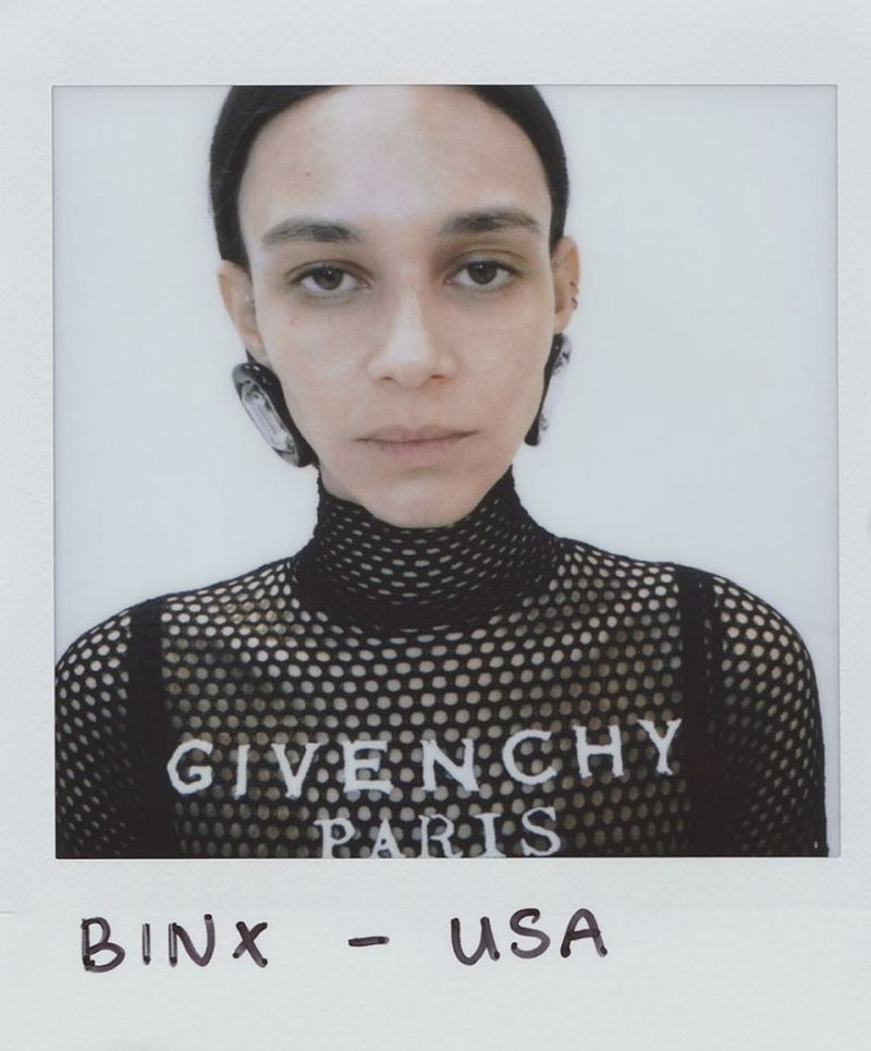 A headshot of a woman with black hair and large clear earrings wearing a black mesh top with “Givenchy Paris” printed on it.