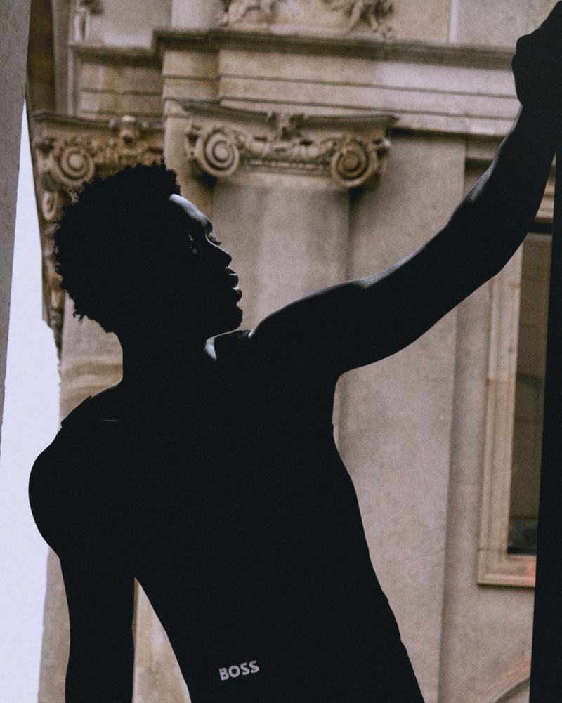 Silhouette of a Black man holding onto a stone pillar, with the Boss logo visible on his garment and classical architecture behind him.
