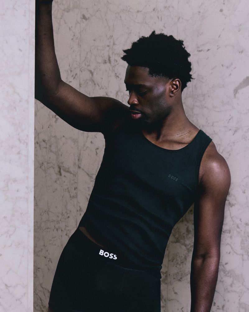 Athletic Black man in a tank top and leggings leans against a marble wall, showcasing sportswear fashion in an indoor environment.