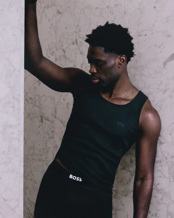 Athletic Black man in a tank top and leggings leans against a marble wall, showcasing sportswear fashion in an indoor environment.