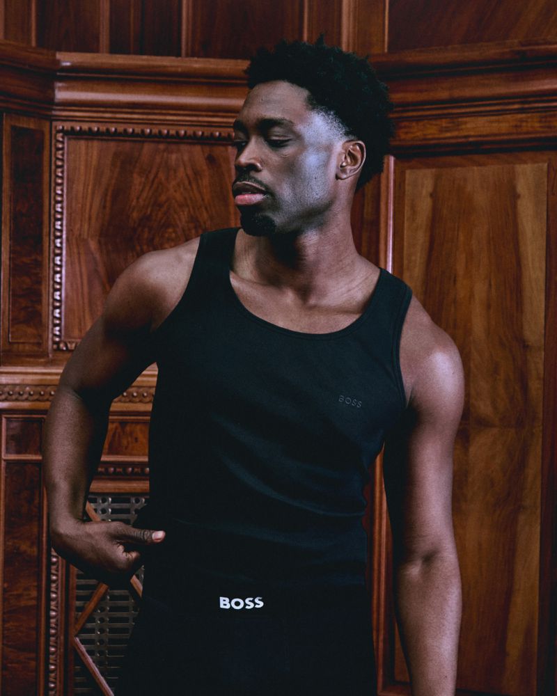 Athletic African American man poses in a black tank top and pants, highlighting his physique, against a wood-paneled wall indoors.