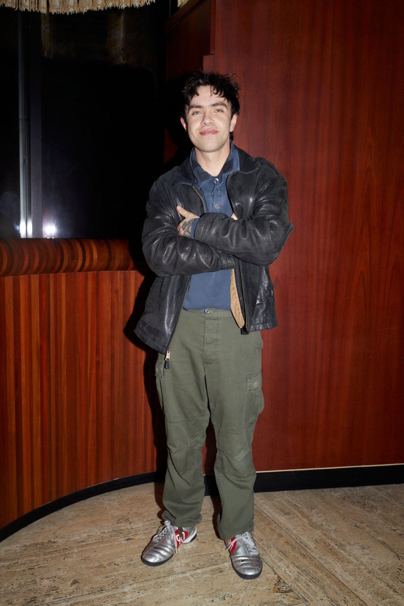 Full body shot of a smiling young adult male with his arms crossed, wearing a black leather jacket, blue shirt, and green pants.