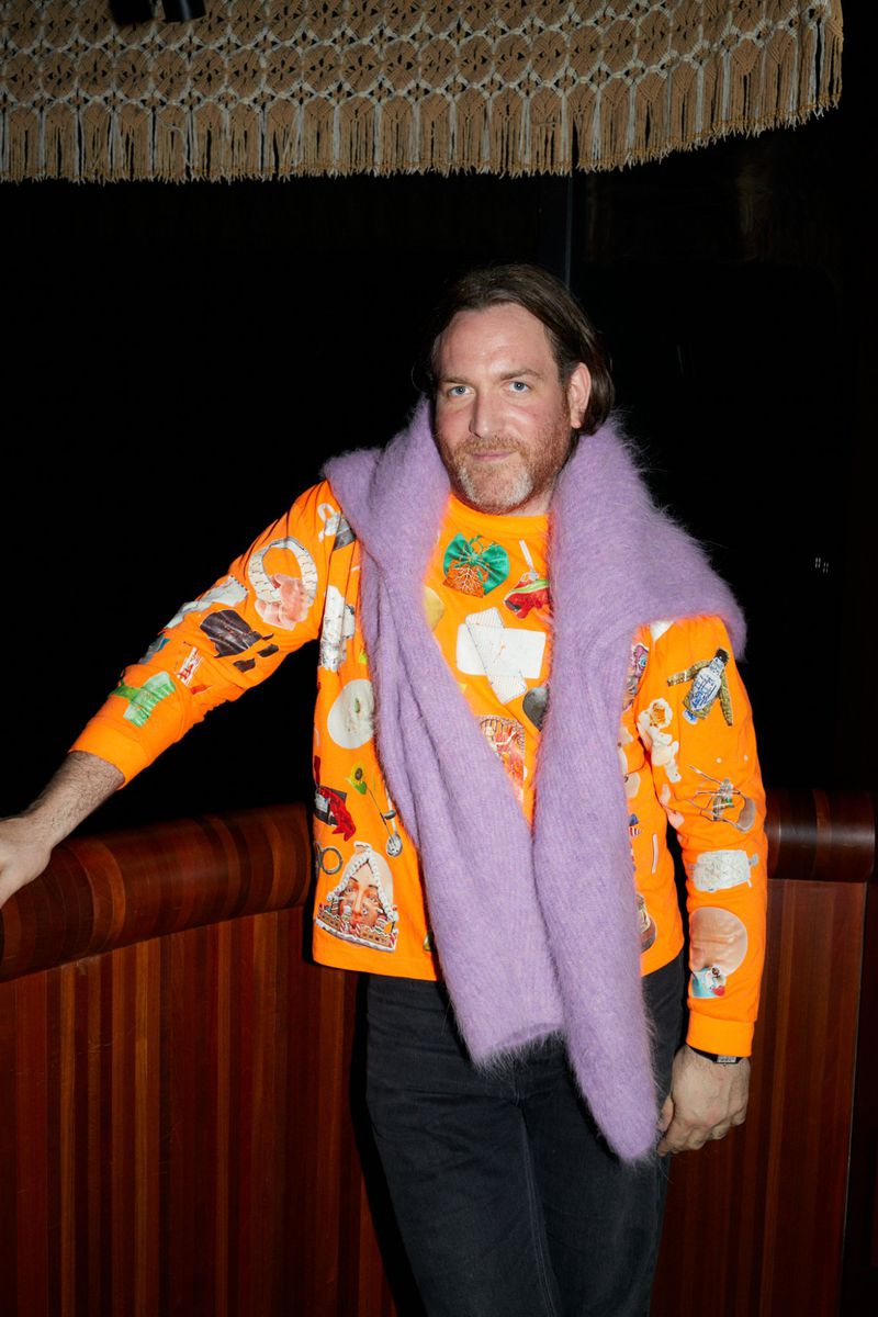 Adult man posing indoors wearing a patterned orange sweater, light purple scarf, and dark pants in front of a brown wood wall.