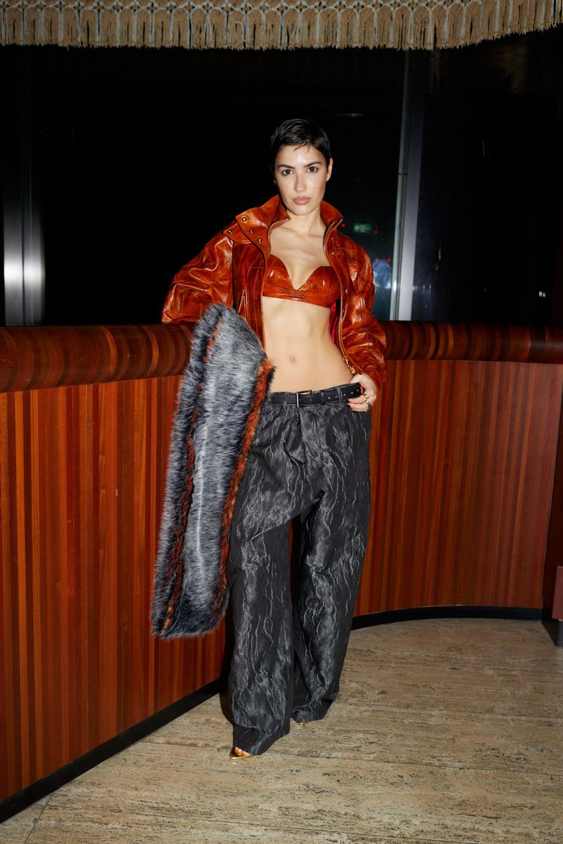 Adult female in a faux-fur-lined jacket and bra top poses indoors with a pair of high-waisted, dark-colored slacks and a belt.