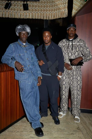 Three men stand indoors, showcasing fashion; one wears a denim outfit, another in navy, and the third in a snakeskin-patterned suit.