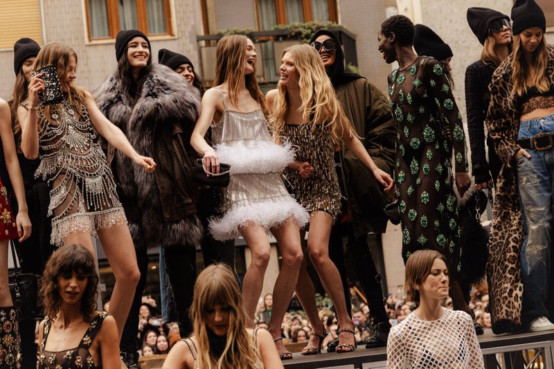 Group of fashion models pose outdoors on a platform wearing dresses, fur coats, and beanies with a crowd visible below.