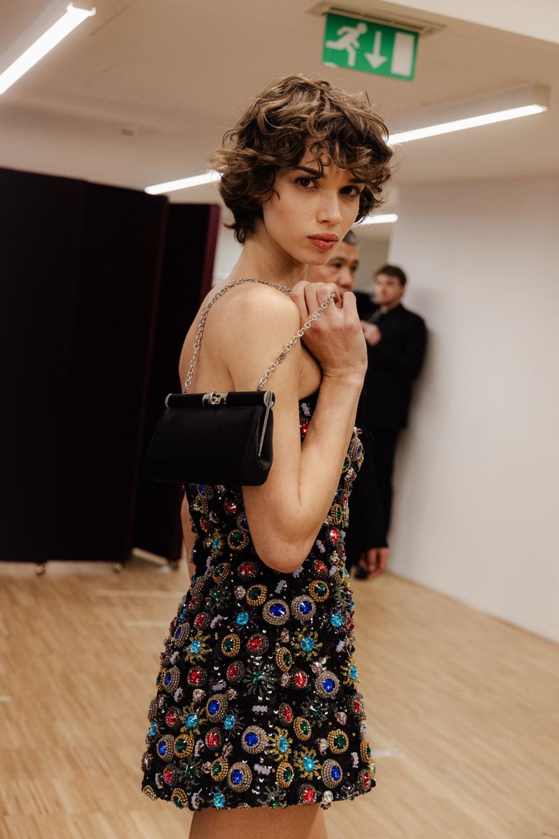 A woman with curly hair poses indoors wearing a beaded mini dress and holding a black purse on a chain, with people blurred in the background.