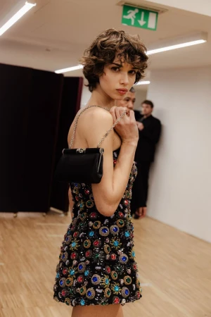 A woman with curly hair poses indoors wearing a beaded mini dress and holding a black purse on a chain, with people blurred in the background.