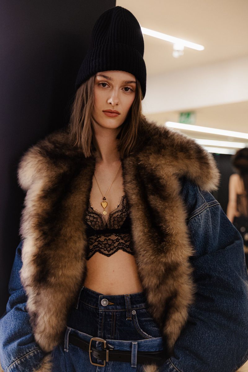 A woman poses indoors wearing a black beanie, fur-trimmed jacket, lace top, and jeans, gazing directly at the camera with neutral expression.