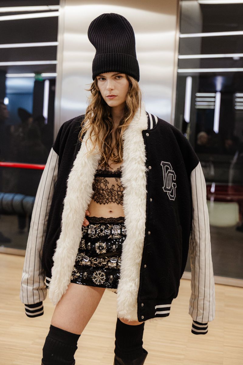 Fashionable woman in a black bomber jacket, sequined skirt, and knit beanie poses indoors; chic urban style, confident pose, and trendy fashion.