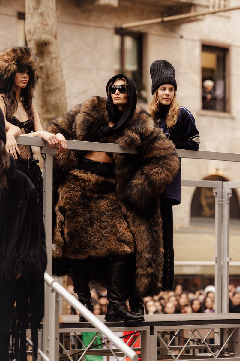 Fashion show models on an outdoor runway wearing statement fur coats, black tops, and black bottoms on a gray, sunny day.