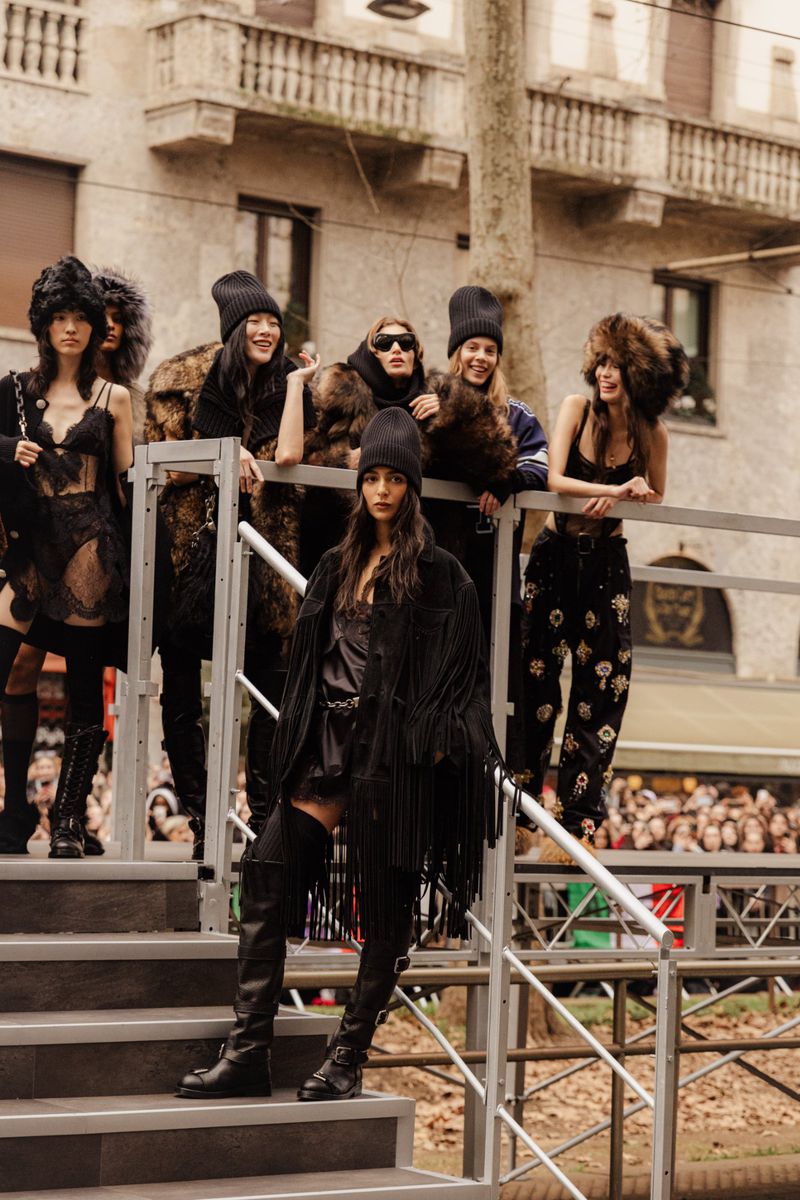 A group of models standing on an outdoor staircase, posing in black clothing with fur hats, with a blurred crowd and building in background.