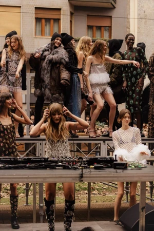 A group of models present a fashion show on a stage with audio equipment on a city street during daylight in an outdoor setting.