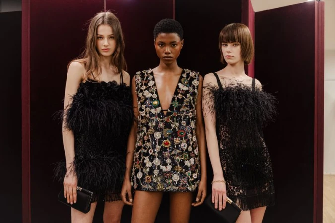 Three models wearing luxury black dresses posing against a burgundy room divider screen
