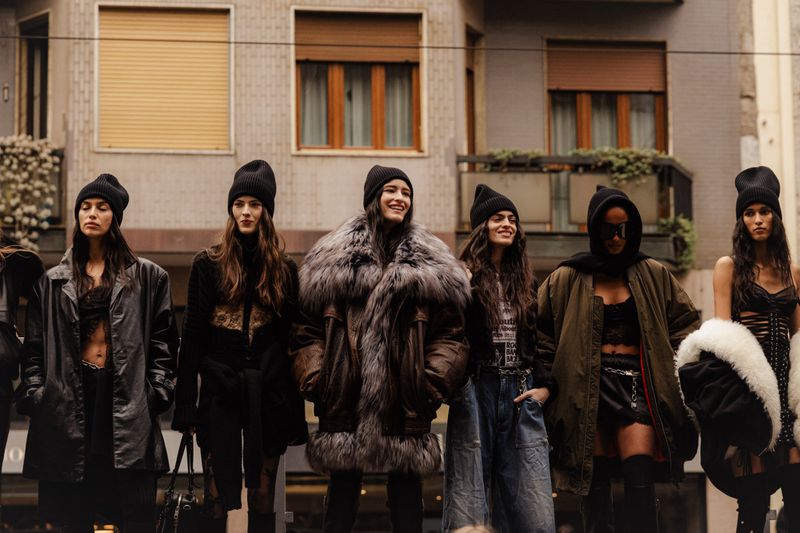 A group of fashion models stand outdoors modeling clothing including coats, jackets, jeans and tops against a neutral urban backdrop.
