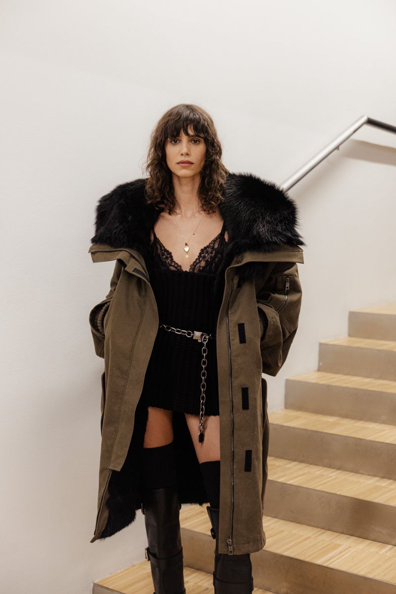 A fashionable woman in a long green coat with a fur collar and black boots poses on a staircase with a white wall.