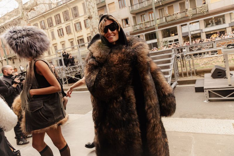 Fashionable adult woman in a fur coat and sunglasses walks past a crowd in an outdoor setting, showcasing street style and urban fashion.