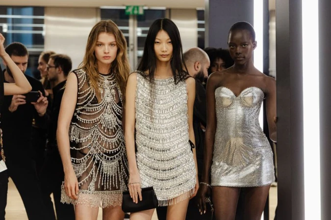 Three diverse fashion models showcase shimmering beaded mini dresses at a fashion show. Elegant indoor setting with a crowd of spectators.