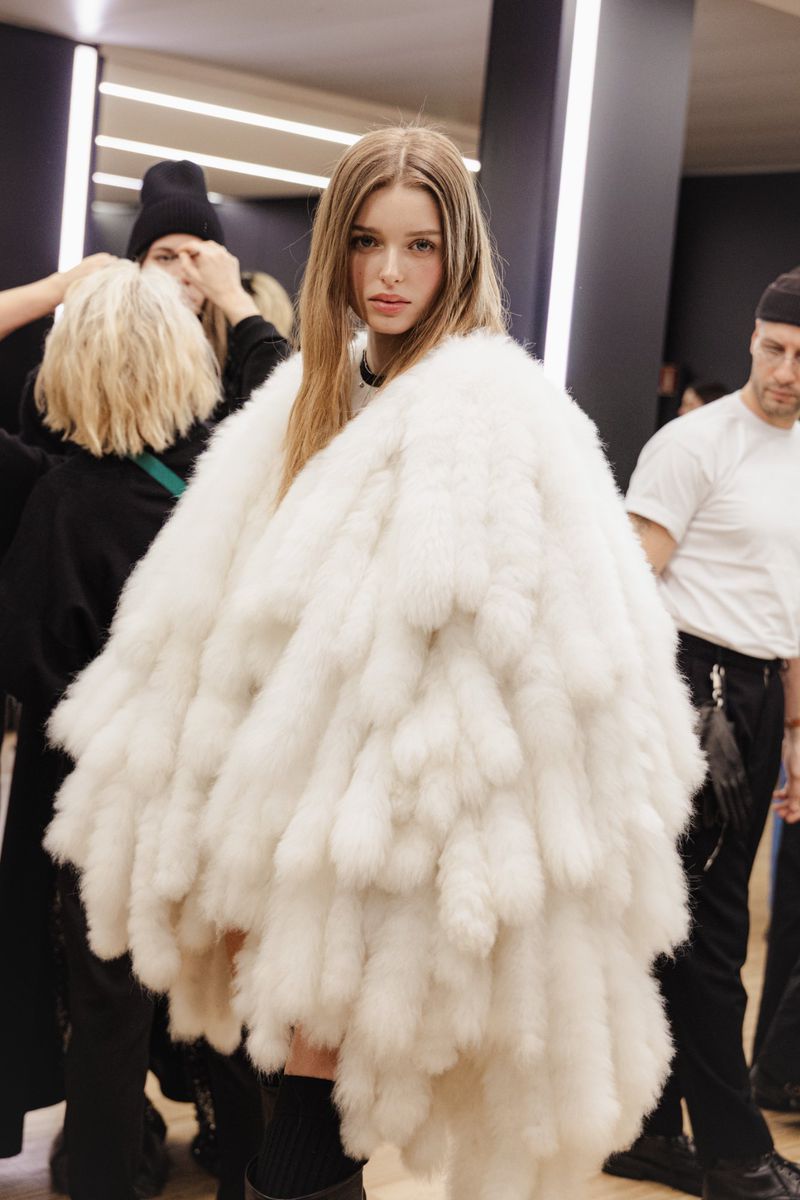 Young woman wearing a large white faux fur jacket stands in an indoor environment with people in the background preparing for a photoshoot.
