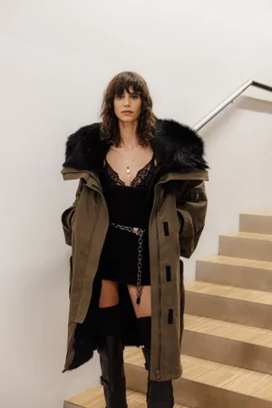 Model wearing an olive green parka with a black fur collar and black boots standing near wooden stairs