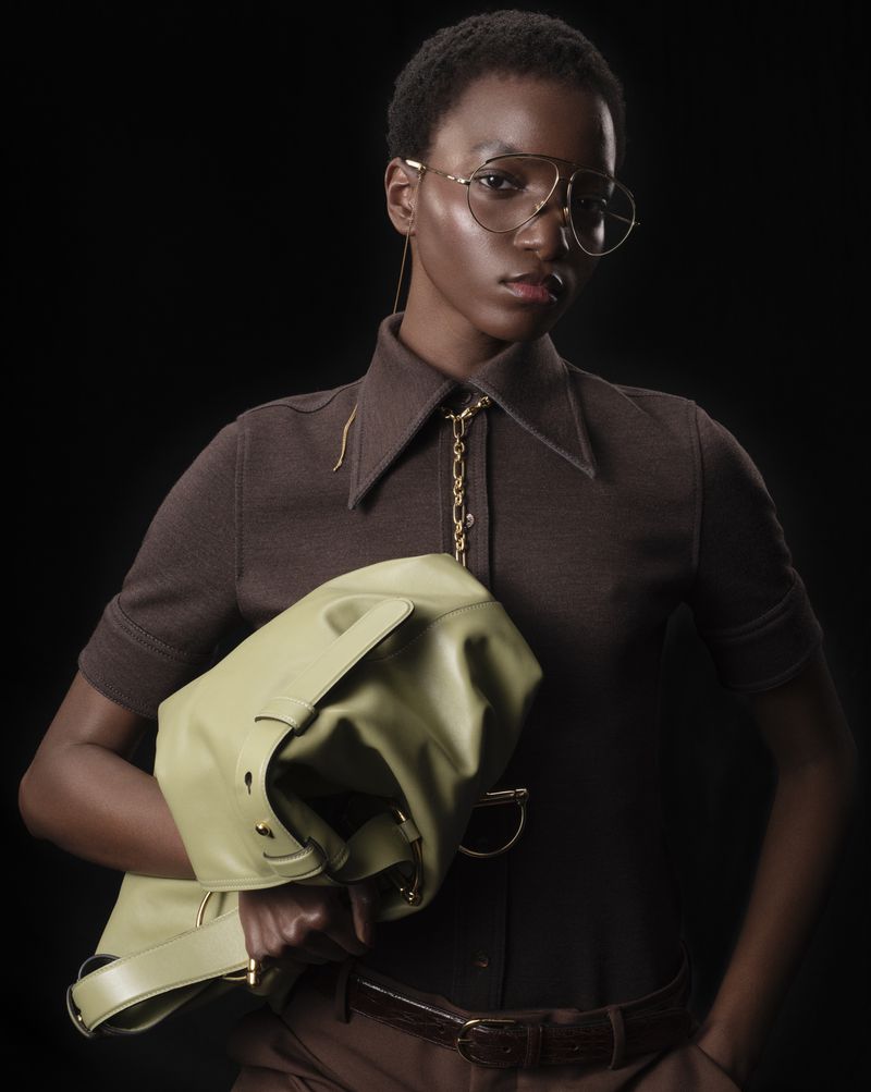 Woman with short, dark hair poses with a light-green leather handbag. She wears glasses and a brown shirt with matching pants against black.
