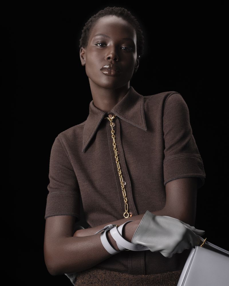 Portrait of a Black woman in a brown collared shirt with a gold chain, wearing grey gloves and holding a grey purse against black.