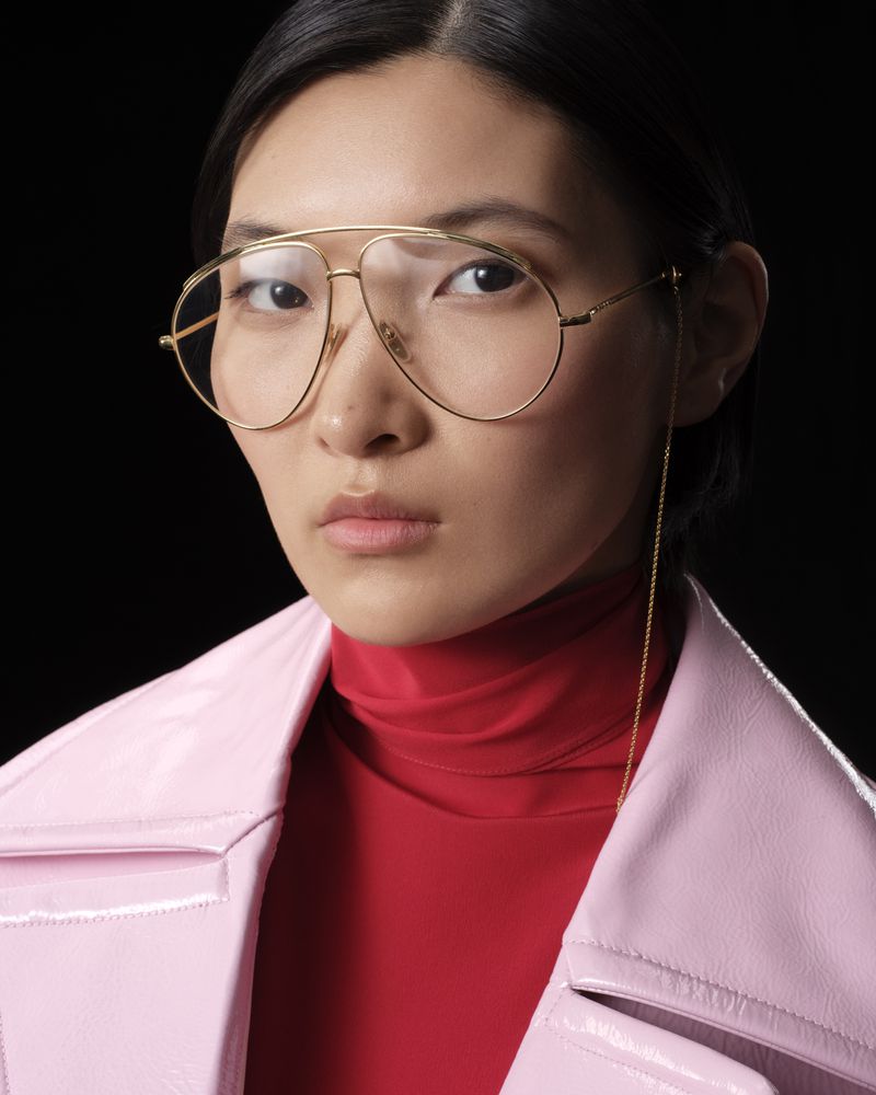 A stylish Asian woman with gold-rimmed aviator glasses, red turtleneck, and pink coat poses against a black backdrop in a close-up.