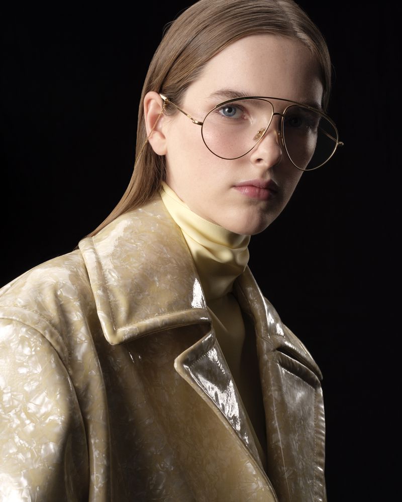 Woman in a beige patent leather trench coat, golden aviator glasses, and a pale-yellow turtleneck against a black background.