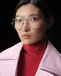 Close-up portrait of a woman wearing gold aviator glasses, a red turtleneck, and a glossy pink coat against a black background.