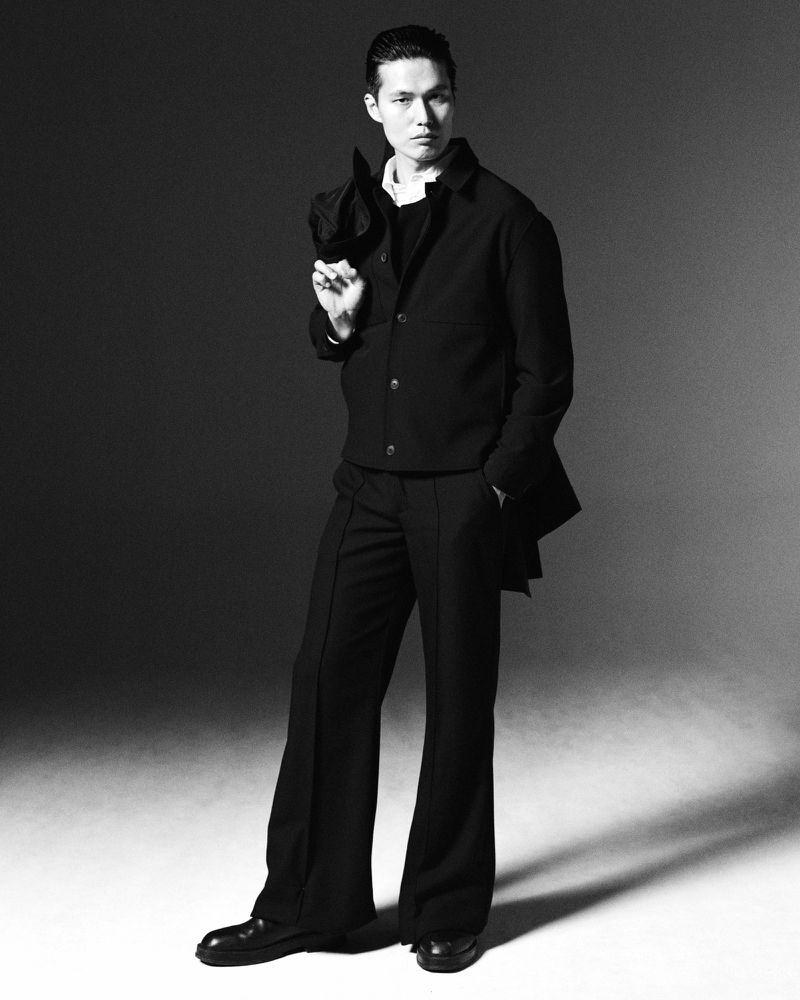 Studio shot of an Asian man in a black jacket and flared pants, holding a coat over his shoulder, against a grey backdrop.