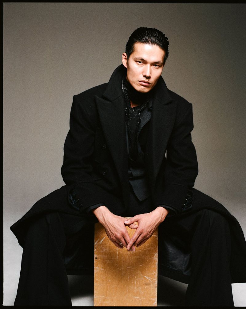 Asian man seated on a wooden box wearing a black coat in a studio setting