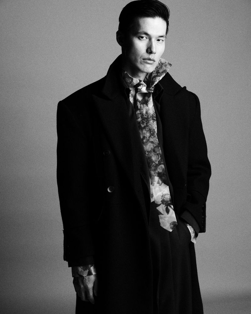 Asian man in black overcoat and floral shirt poses in a monochrome portrait, blending fashion and classic style against a neutral background.