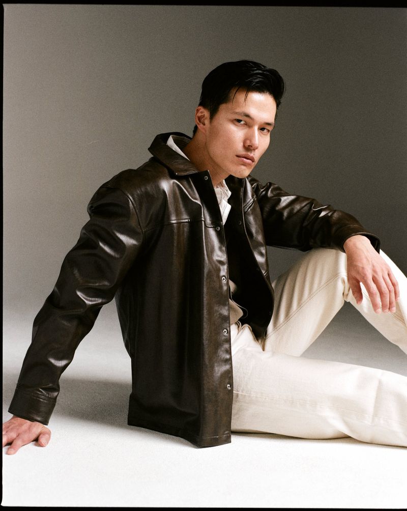 A dark-haired adult male poses indoors, wearing a brown leather jacket and off-white pants, sitting against a neutral grey background.