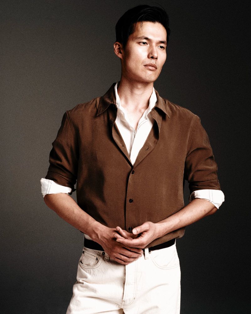 Portrait of a handsome adult Asian man in a brown button-down shirt and white pants against a solid grey background, captured in a medium shot.